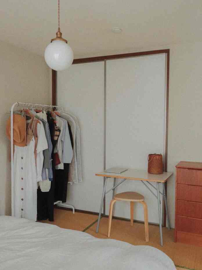 A narrow single household room with white walls and tatami flooring featuring a clothes rack, minimal desk, and brown dresser in a natural tone.