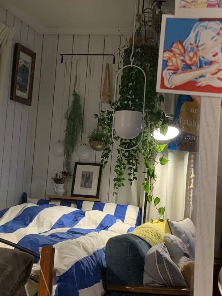 白と青のナチュラルな寝室、中央左に大きなベッドと多様な植物が配置された居心地の良い空間