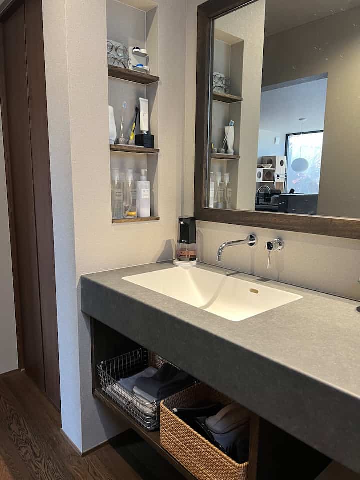 グレーとナチュラルトーンのバスルーム、コンパクトな洗面台、バスケット収納、大きな鏡が配置された落ち着いた空間