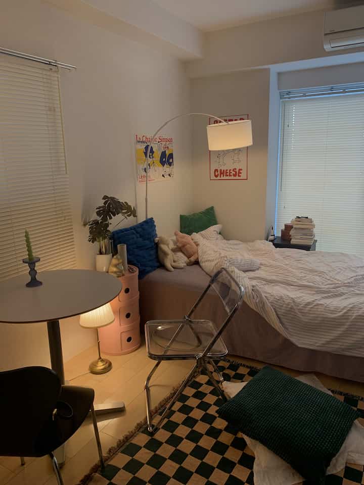 ベージュ色調のワンルーム寝室、ベッドとダイニングテーブル、スカイブルーのクッション、暖かな照明が配置された雰囲気のある空間