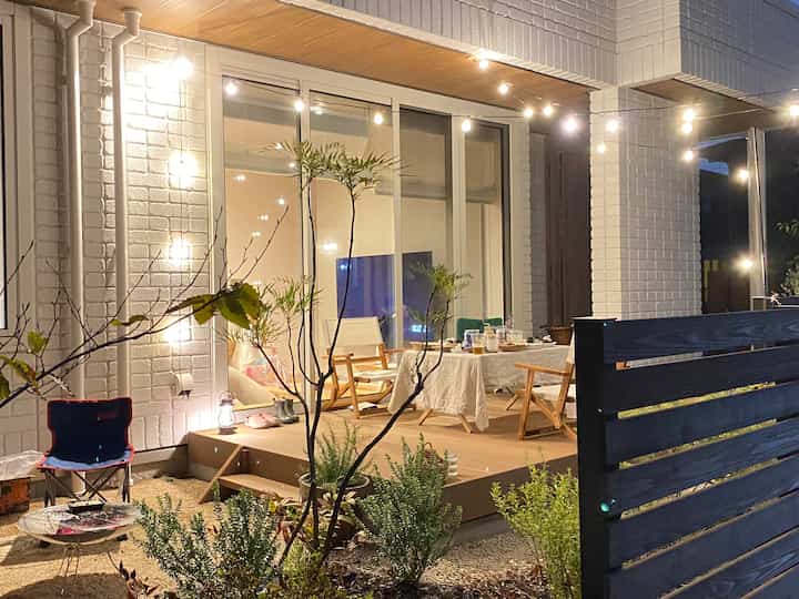 白い壁と茶色のデッキがある小さな庭、ストリングライトの下に配置されたアウトドアテーブルとチェアが特徴的な雰囲気のあるインテリア