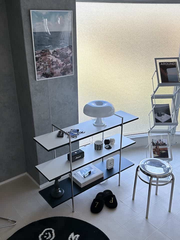 White and silver toned living room featuring a modern stool, shelving unit, and poster creating a clean, sleek atmosphere