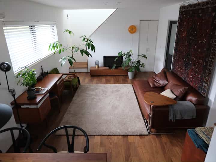 木目調と白い壁のナチュラルモダンなリビング、ブラウンレザーソファとデスク、植物が配置された落ち着いた空間