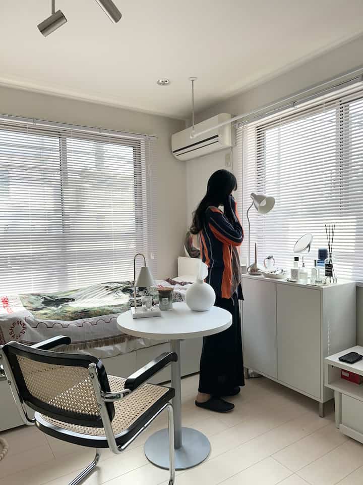 白いトーンの寝室兼リビング、ブラインドと丸テーブル、シンプルな家具が配置された落ち着いた雰囲気のインテリア