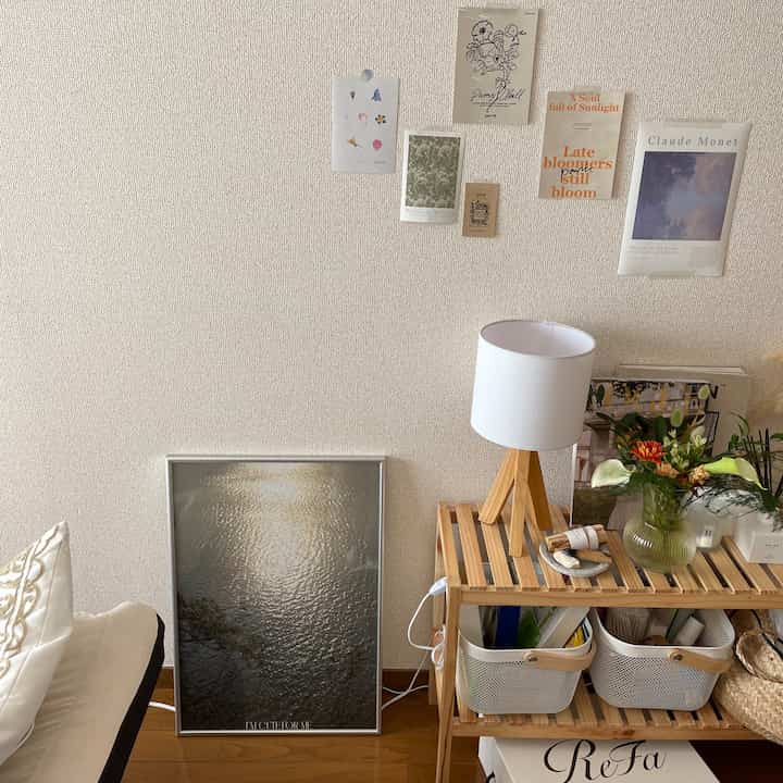 白い壁とナチュラルな木目調の棚があるリビング空間、シルバーフレームのポスターとフロアランプが落ち着いた雰囲気を演出。