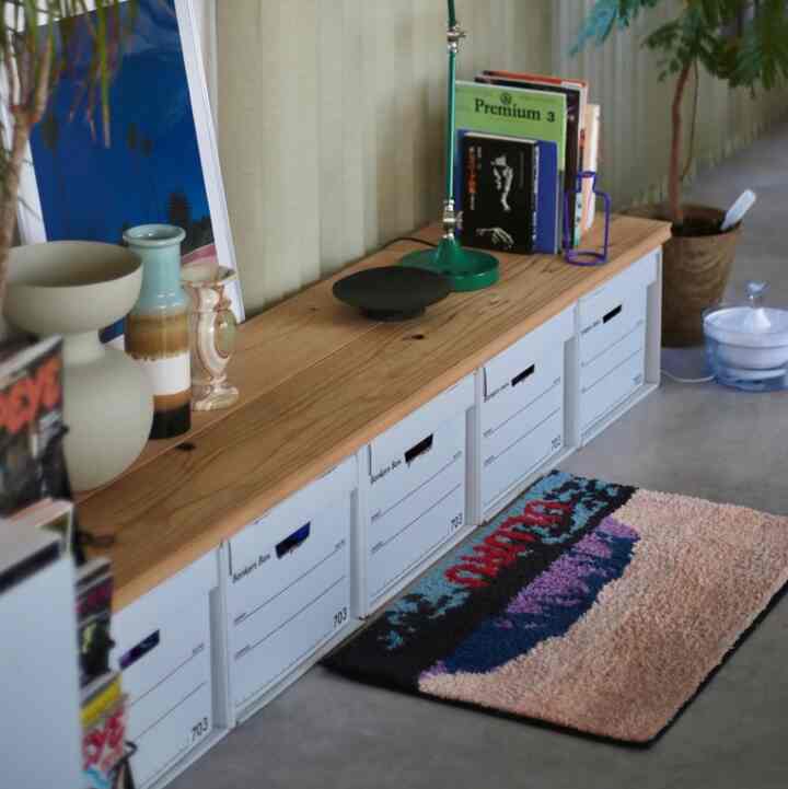 ナチュラルモダンな雰囲気の、木目調のローキャビネットと白い収納ボックスが配置され、カラフルなラグと植物が置かれた印象的な空間