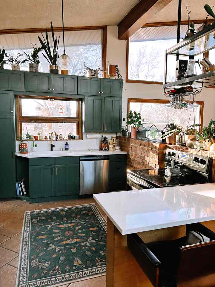 Warm kitchen space with green cabinets and natural light, featuring plants and a patterned rug