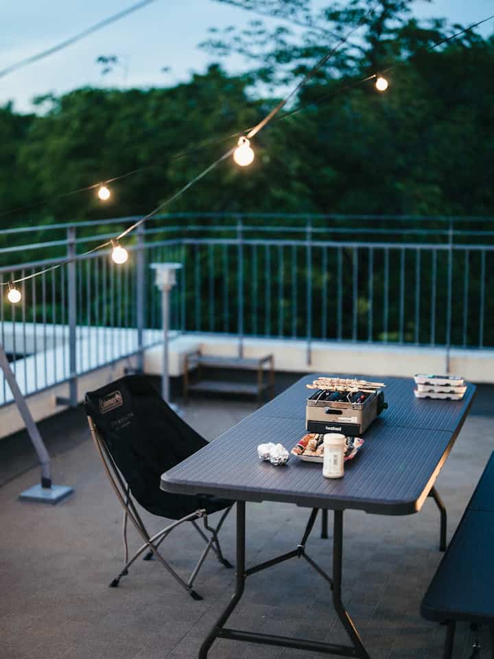 Cozy terrace balcony with green foliage backdrop, metal railing, outdoor table and folding chair arranged for an inviting evening