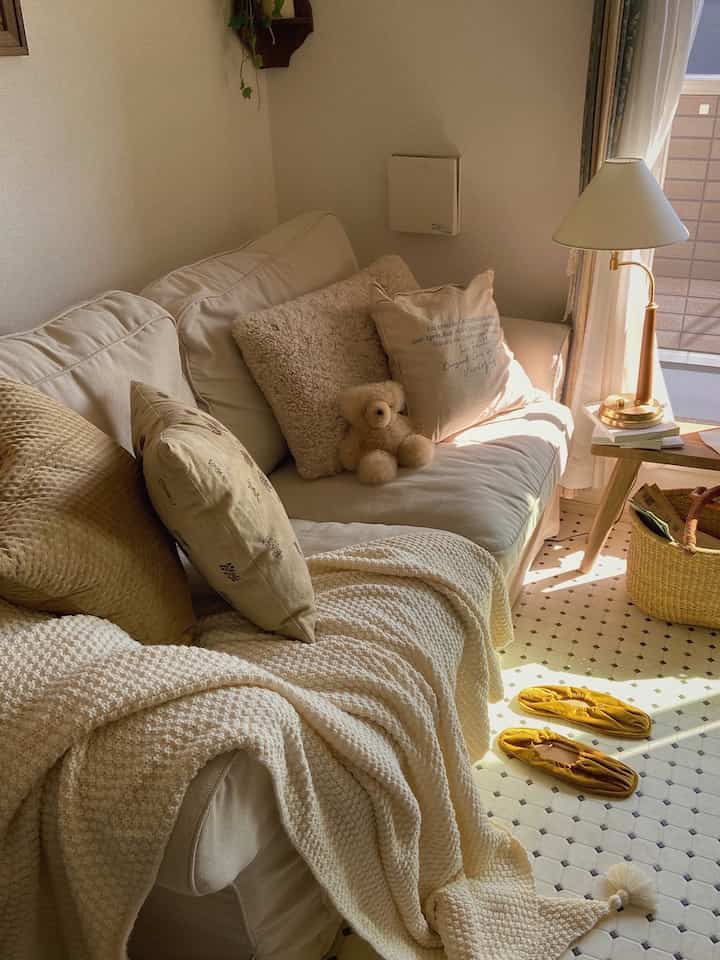 アイボリーとベージュの色調のリビング、ソファにブラウンのクッションとブランケットが配置された陽光が差し込む雰囲気のあるインテリア