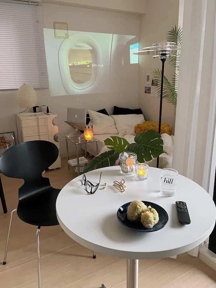 Modern studio living room with white walls and light wood floor, featuring black armchair, transparent acrylic side table, and projector screen