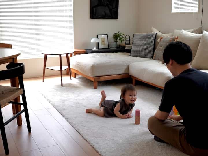 明るい木目調と白が調和するリビング、ベージュのソファと大きなラグの上で赤ちゃんと大人が一緒に過ごす雰囲気のある空間