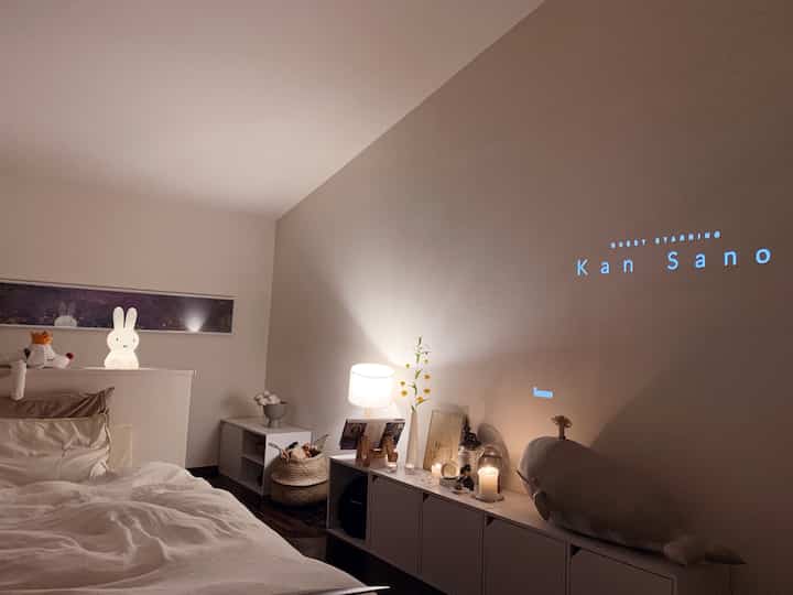 White and natural tone bedroom featuring bed, modern cabinet topped with lighting and a projected video creating a cozy atmosphere