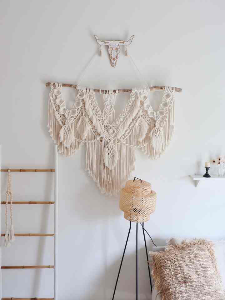白とベージュの色調のリビング、マクラメの壁飾りとフロアランプ、クッションが配置されたナチュラルで雰囲気のあるインテリア