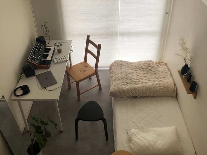 ナチュラルカラーのコンパクトな一人暮らし寝室、白いデスクと木製の椅子、ベージュの寝具が配置されたミニマルなインテリア