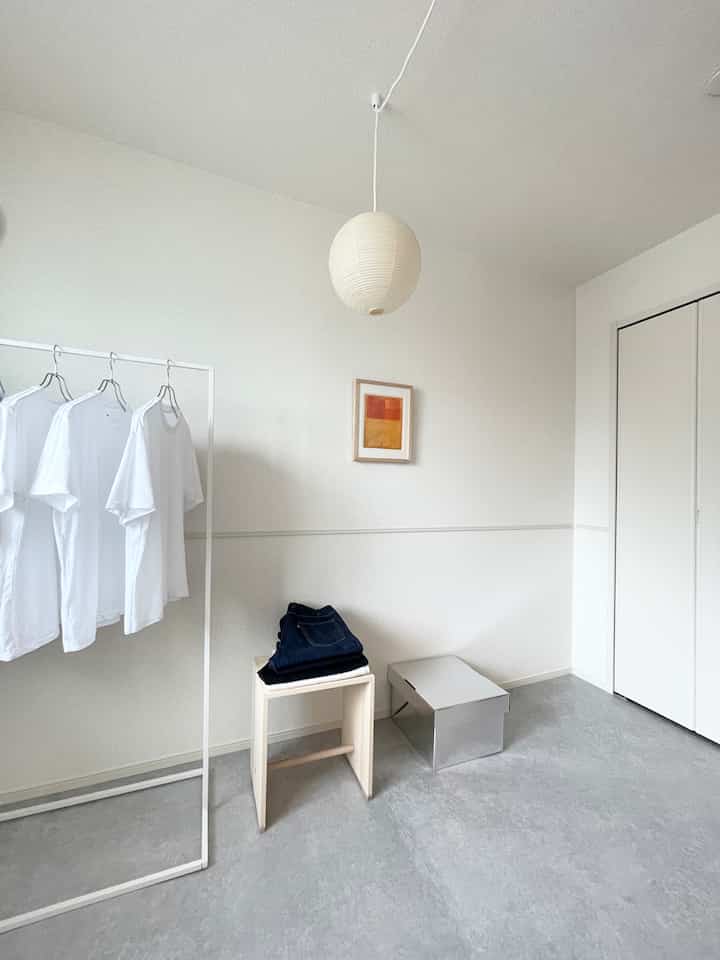 Minimal white-toned living room featuring a clothes rack, stool, and archive box with a clean, spacious atmosphere