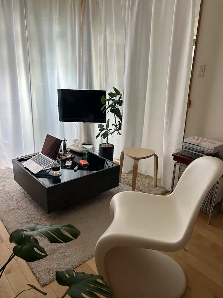 White-curtained living room featuring a mid-century modern chair, black coffee table, and plants creating a cozy atmosphere