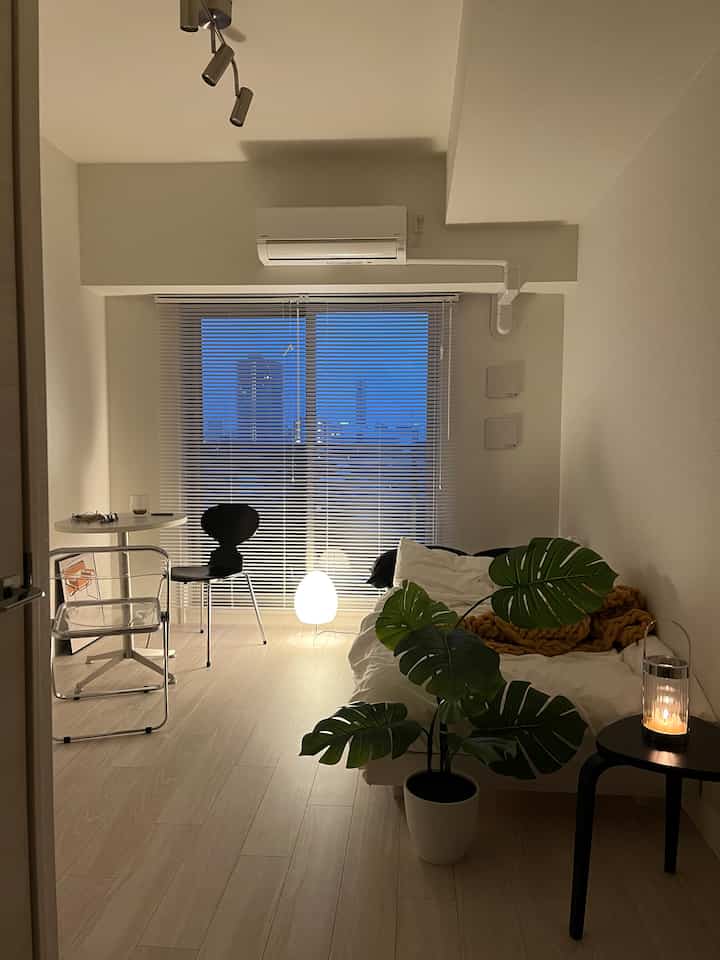 A white and wood tone 8-tatami L-shaped studio room featuring black chairs, a Monstera fake plant, cozy lighting, and minimalist decor