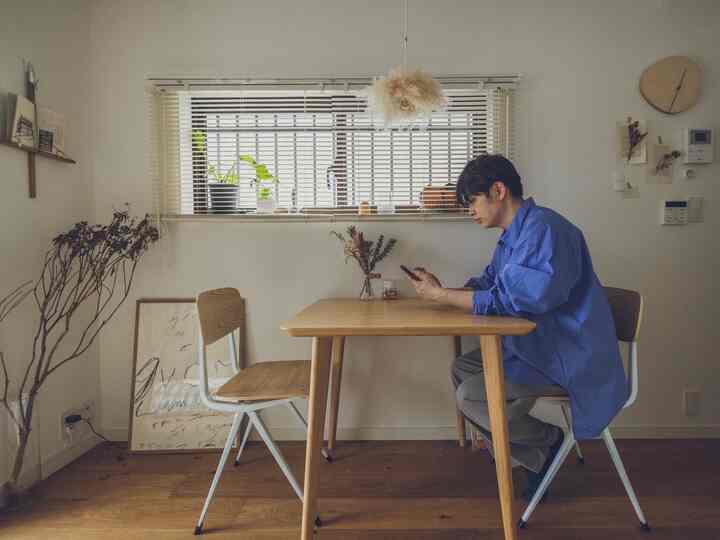 ナチュラルな木目調と白い壁の8畳ダイニングルーム、中央に木製の食卓と椅子二脚、自然光の下で携帯電話を使う男性の様子