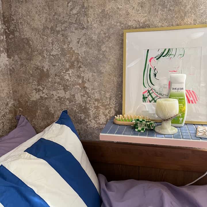 Gray-beige concrete wallpaper bedroom with natural wooden bed frame, blue striped pillow, and nightstand featuring candle and decor items for a cozy feel