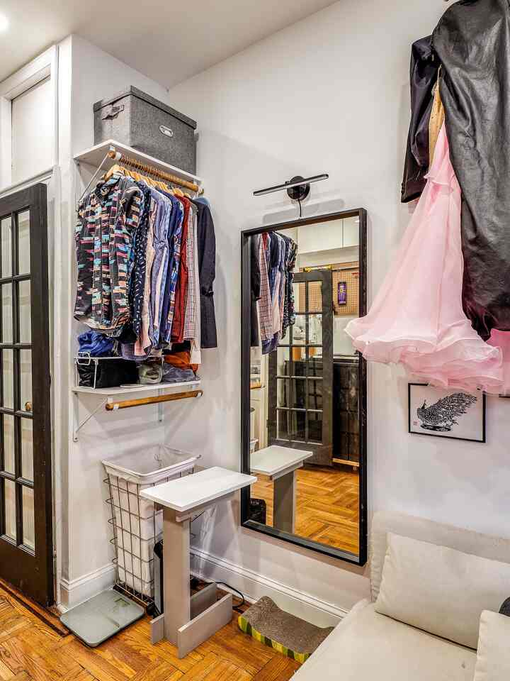 White walls and light brown floor define a narrow dressing room with wall-mounted closet shelves and a black-framed mirror