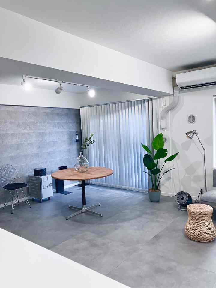 グレーのコンクリート壁と床、丸い木製のダイニングテーブル、ラタンのスツール、緑の植物が配置されたモダンなインダストリアルキッチンスペース