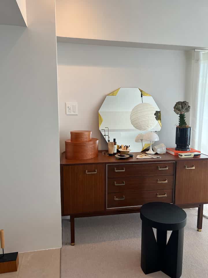 Brown wood tone sideboard against white walls in living room, featuring an octagonal mirror, cosmetics, and black stool creating a natural atmosphere