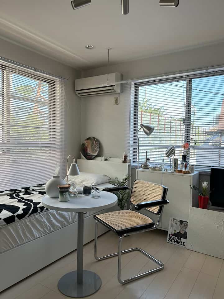White and natural toned bedroom featuring a small table by the window and a natural material chair in a calm space