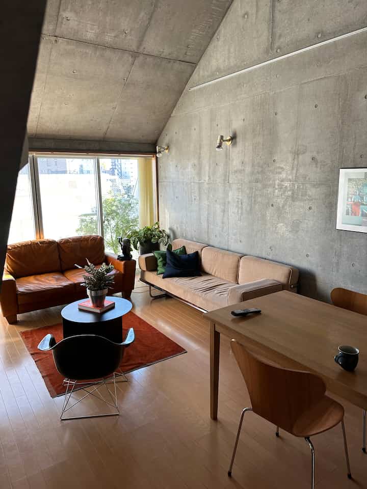 コンクリート打ちっぱなしの壁と木目調の床が調和したミッドセンチュリーモダンのリビングルーム、レザーソファとベージュソファが配置された落ち着いた空間