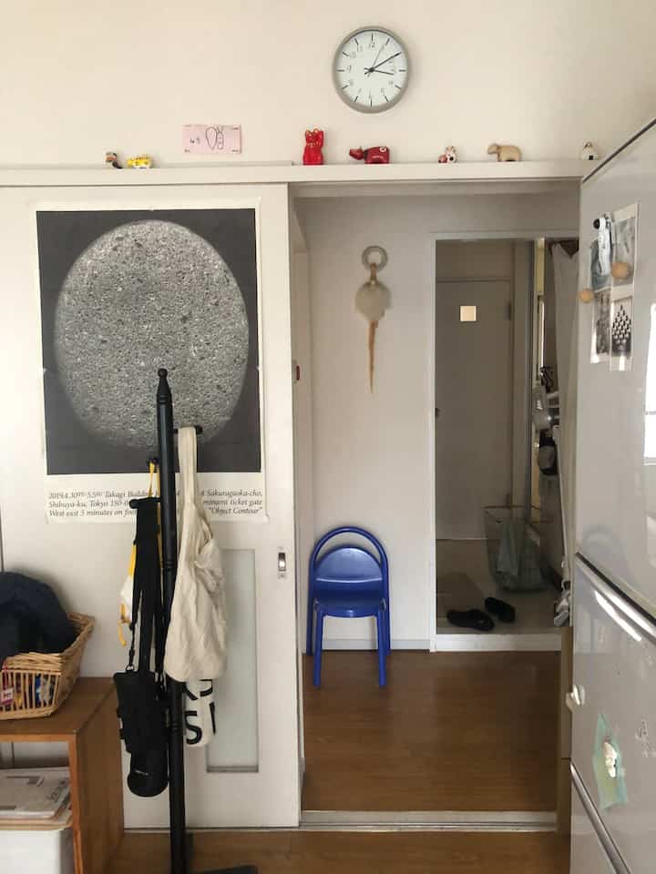 白い壁と茶色の木の床が調和する狭い玄関スペース、青い椅子と壁の時計がアクセントとして配置されている