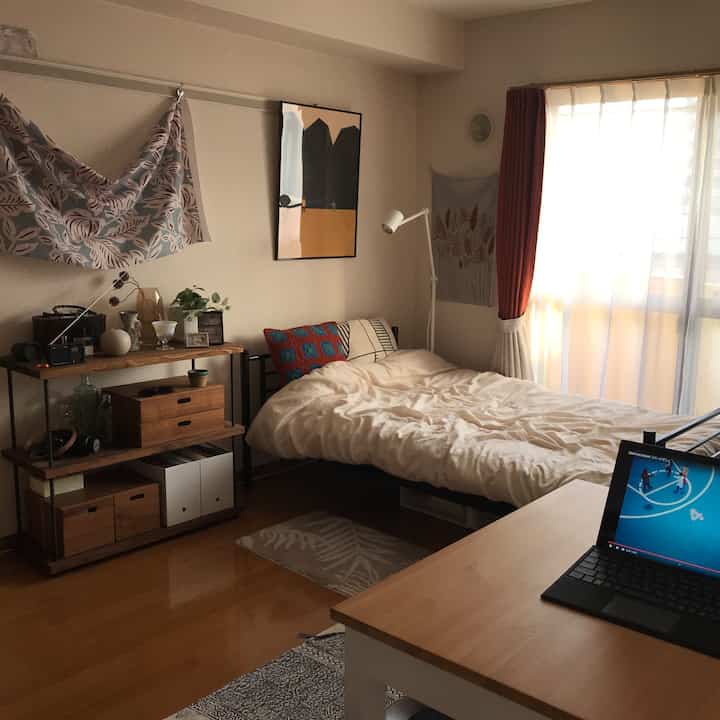 A simple single-person bedroom with white walls and wood-tone furniture, featuring a desk with a laptop and floor lamp for a cozy atmosphere