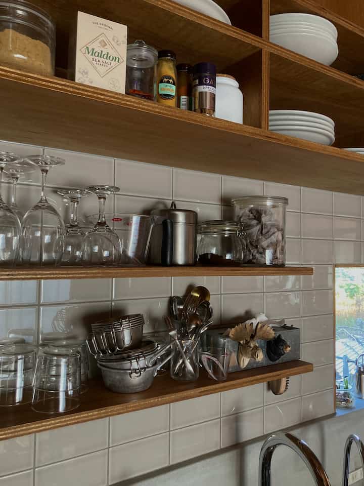 白いタイルの壁と木目調の棚に調理器具や皿、グラスが整えられたキッチンの雰囲気あるインテリア