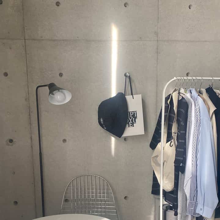 グレーのコンクリート壁に白い衣類ラックと黒のフロアランプが配置されたミニマルなワンルームの玄関