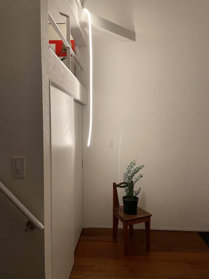 白い壁と茶色の木製床が特徴的な小さな玄関、壁に間接照明のLEDライトがあり、椅子の上に植物が置かれている印象的な空間