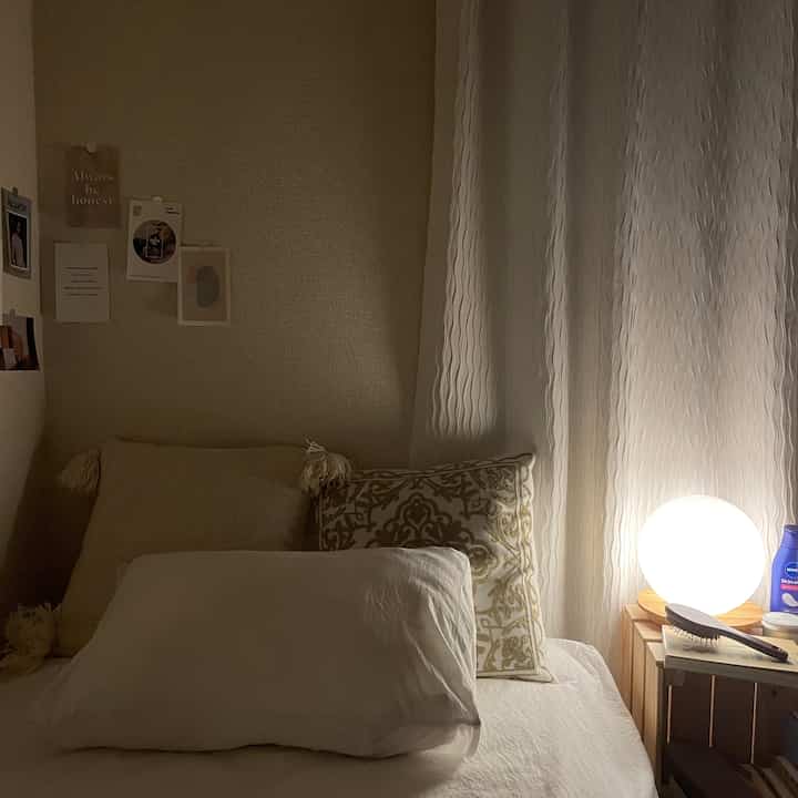 柔らかなベージュ色調の寝室、ベッドとクッション、カーテン、ナイトテーブルとテーブルランプが配置された居心地の良いインテリア