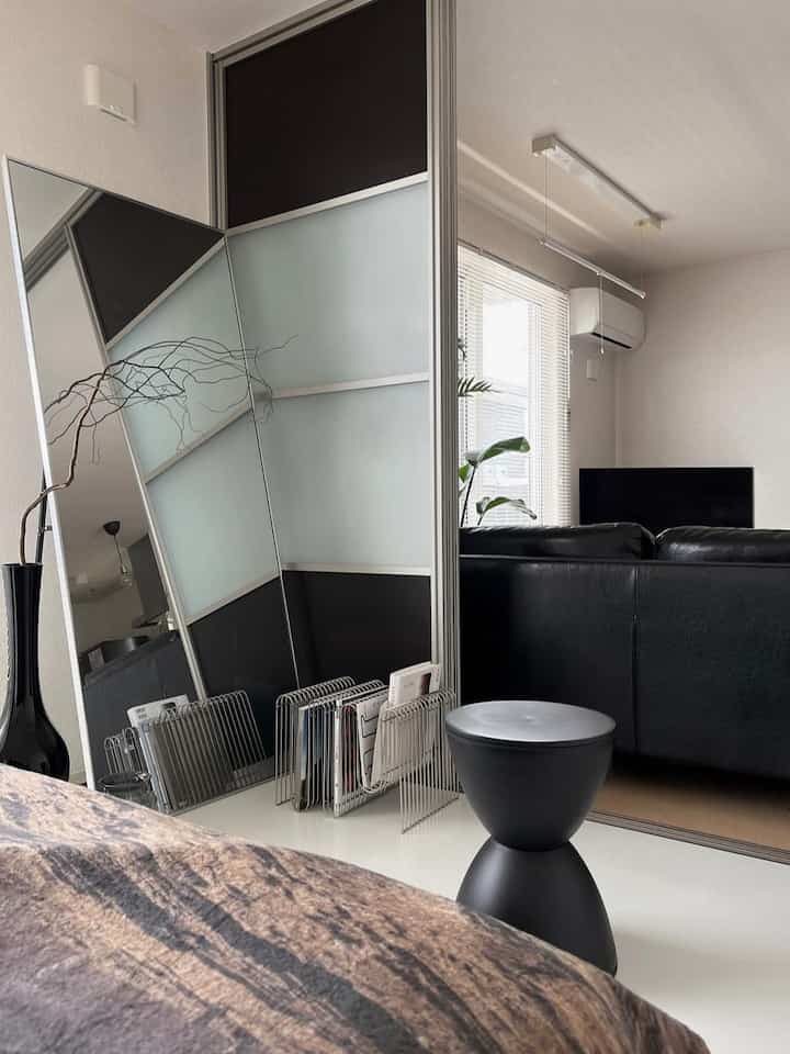 白と黒を基調としたリビング、中央右に黒のソファとスツール、左側に大きな鏡と雑誌ラックが配置されたミニマルな空間