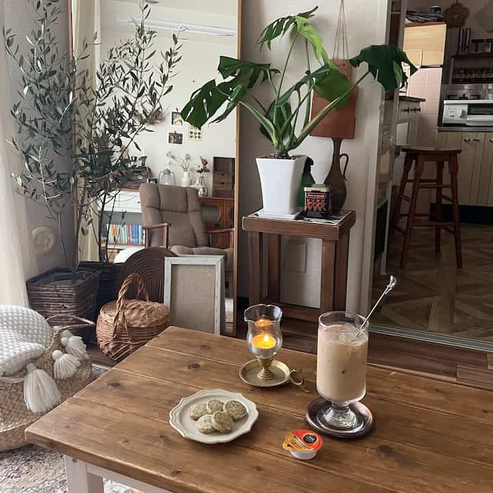 Natural-toned studio living space featuring plants and a wooden dining table with drinks and a candle, creating a cozy home cafe atmosphere