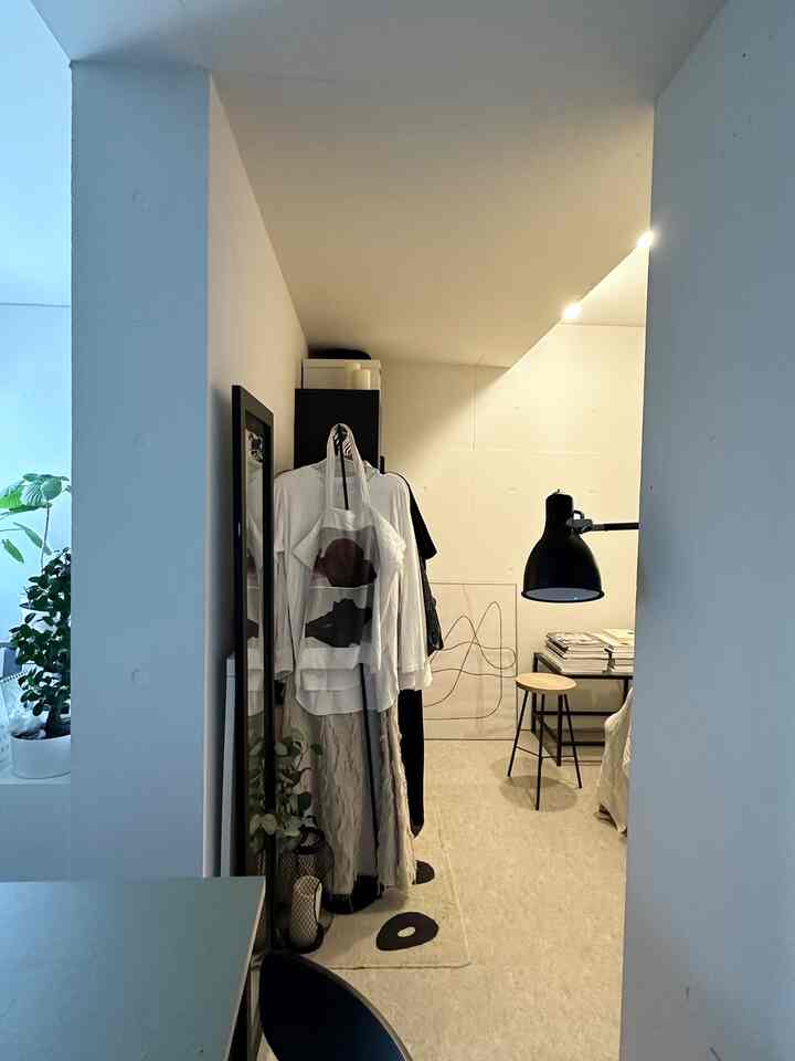 白を基調とした狭い部屋、コーナー収納と黒いフロアランプ、スツールが配置されたミニマルなインテリア