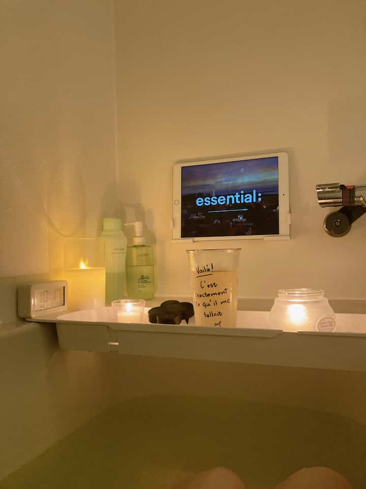 ホワイトトーンのバスルーム、バスタブ上の白いトレイにキャンドルやアロマ洗浄料、飲み物、タブレットが置かれた落ち着いた雰囲気のインテリア