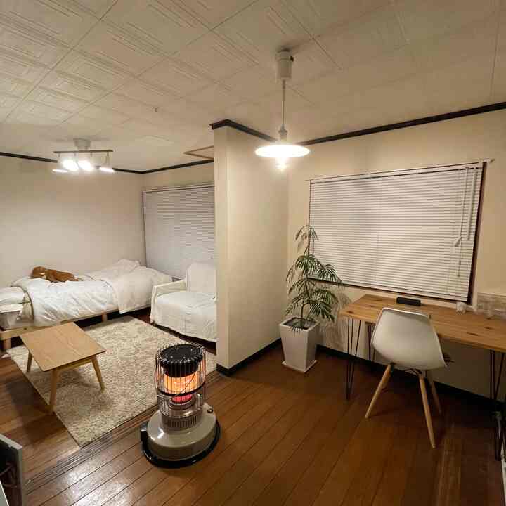 ベージュの壁と木目調の床のコンパクトな1K寝室、白いベッドとデスク、レトロなストーブが暖かい雰囲気を醸し出す室内
