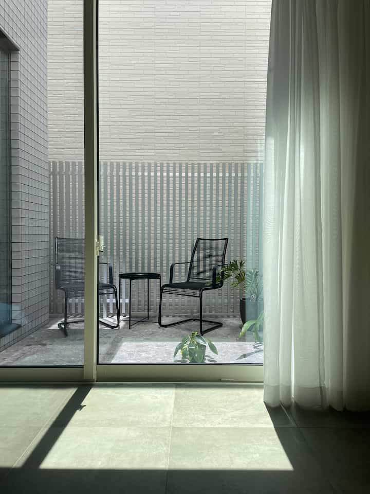 A naturally lit space featuring gray tiles and black outdoor chairs with a side table on a balcony visible through large transparent sliding doors