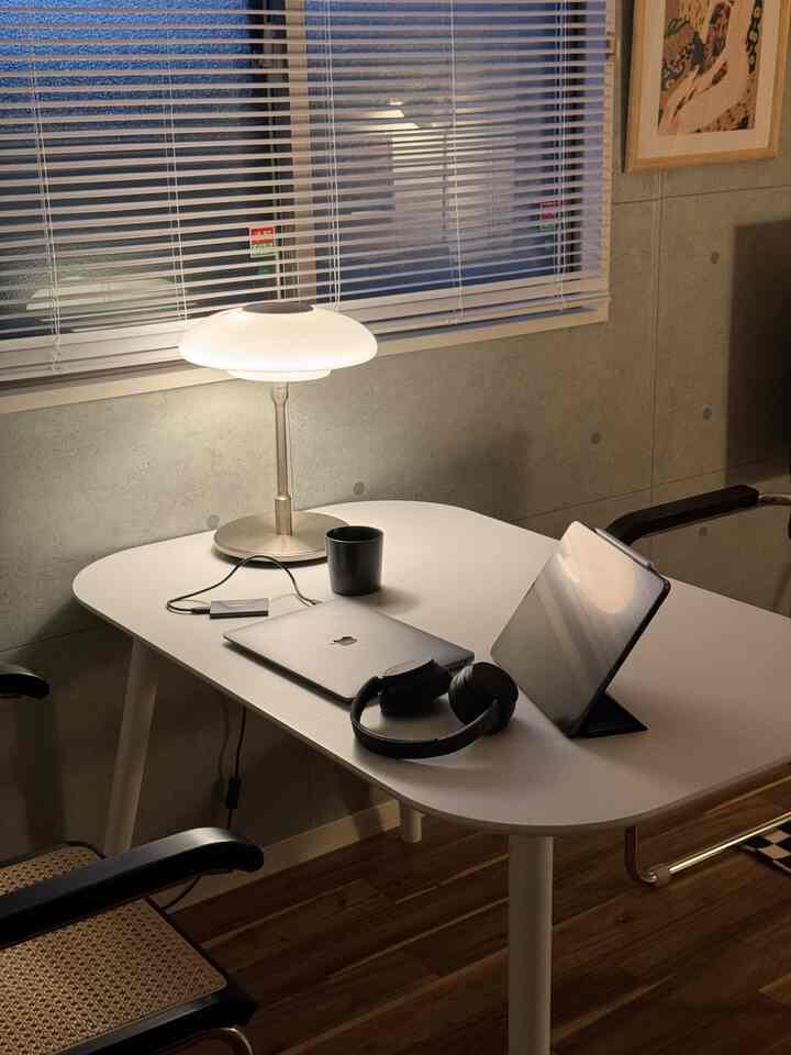 グレーのコンクリート壁と白いダイニングテーブルが配置されたワークスペースのダイニング、テーブルランプとノートパソコンがある落ち着いた雰囲気の空間