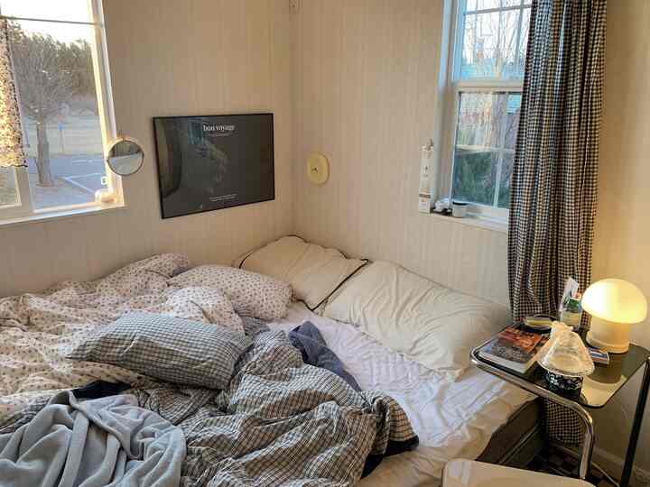 Cozy small bedroom in warm brown and white tones, featuring bed with pillows, a poster on wall, and checkered curtains