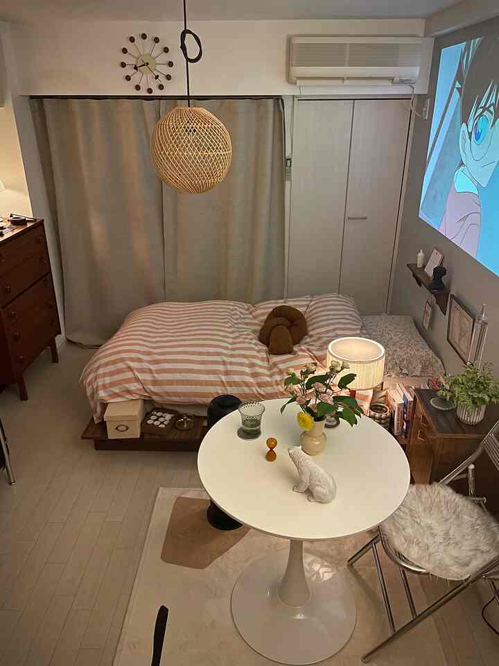 ナチュラルカラーの小さな寝室、ストライプの掛け布団と丸テーブル、温かみのある照明で落ち着いた読書スペースのある雰囲気のインテリア