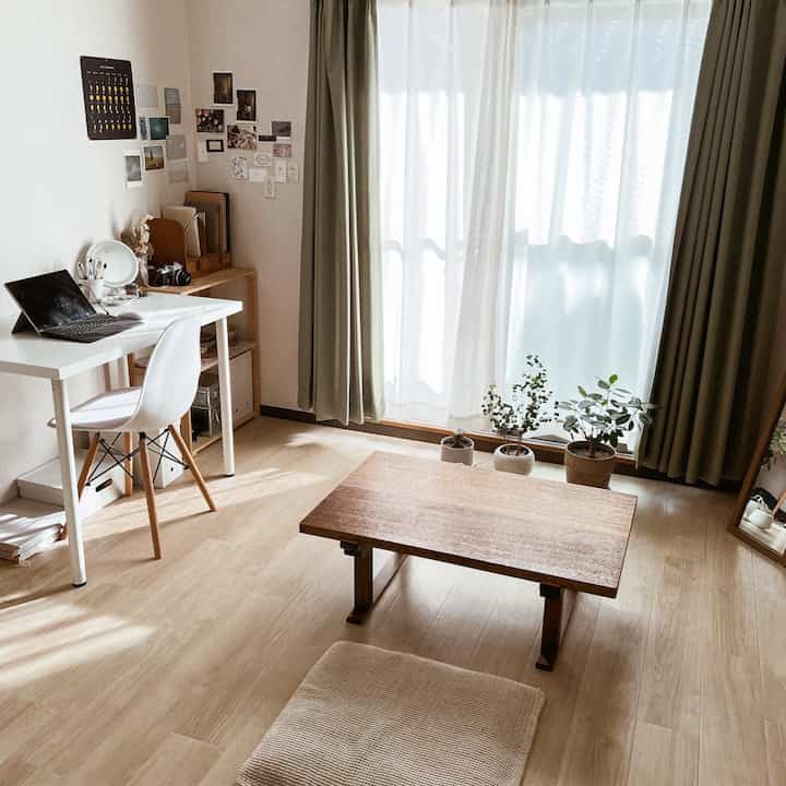 Natural tone studio apartment space featuring white desk, wood-tone low table, curtains, and plants in a cozy home office