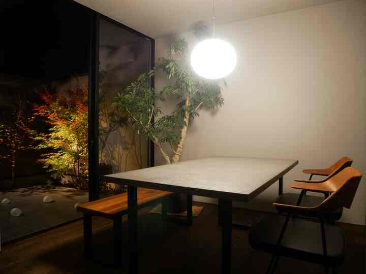 グレーとウッドトーンのダイニングルーム、コンクリートのダイニングテーブルと木製椅子、大きな緑の植物が配置された雰囲気のある空間