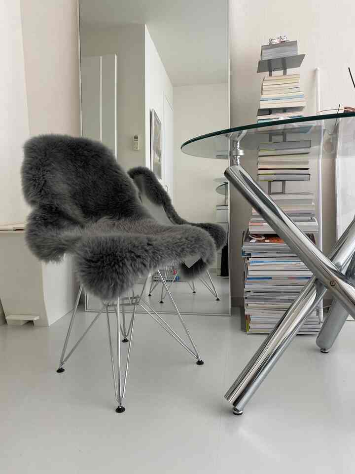 白い壁と床にグレーのシープスキンをかけたダイニングチェアとクローム脚のガラステーブルが配置されたモダンなダイニング空間