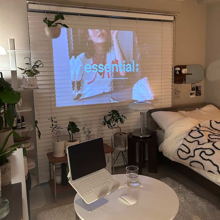 白を基調としたリビング、ブラインドにプロジェクター映像が映され、ベッドと丸テーブルが配置された落ち着いた空間