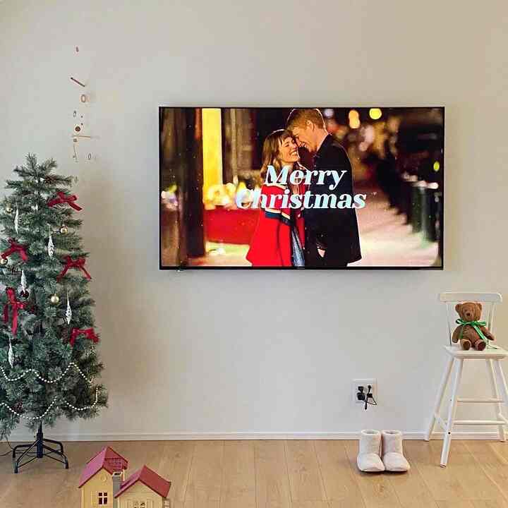 白い壁と木目調の床のリビング、左側にクリスマスツリー、床に小さなおもちゃの家、右側に子供用椅子とテディベアが配置された雰囲気のある空間