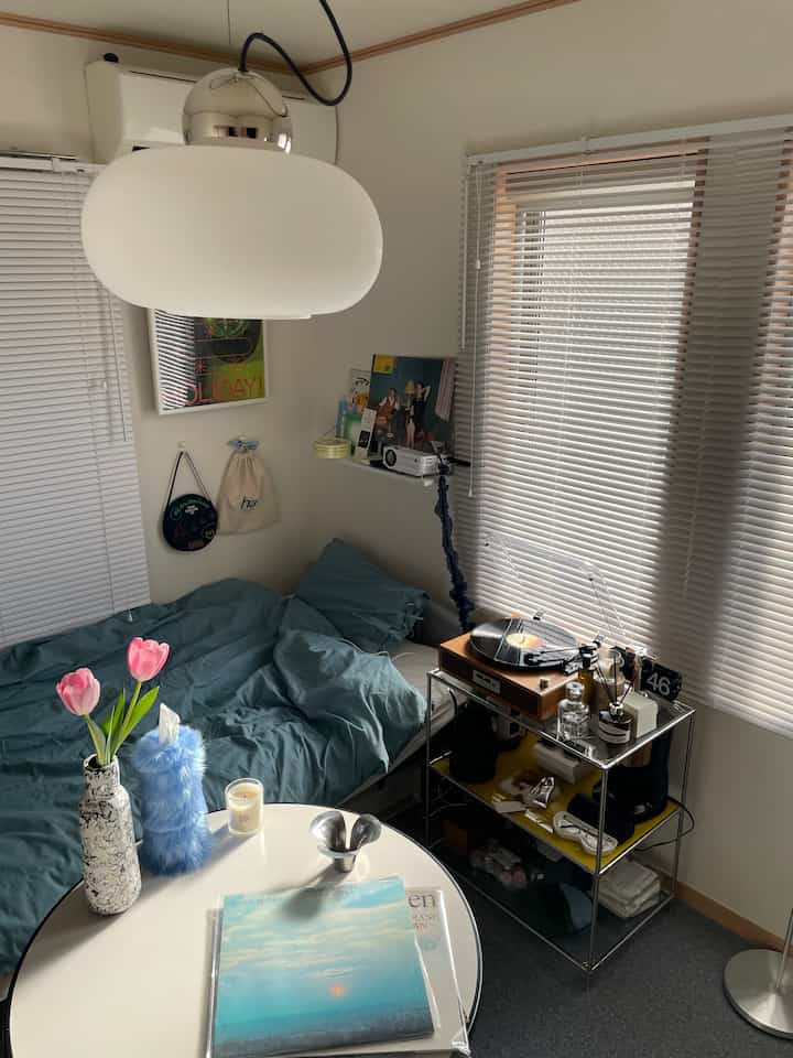 柔らかなブルーのトーンが漂う寝室、中央にペンダントライトとブラインド付きの窓、ベッドとコーヒーカートが整然と配置された雰囲気のあるインテリア