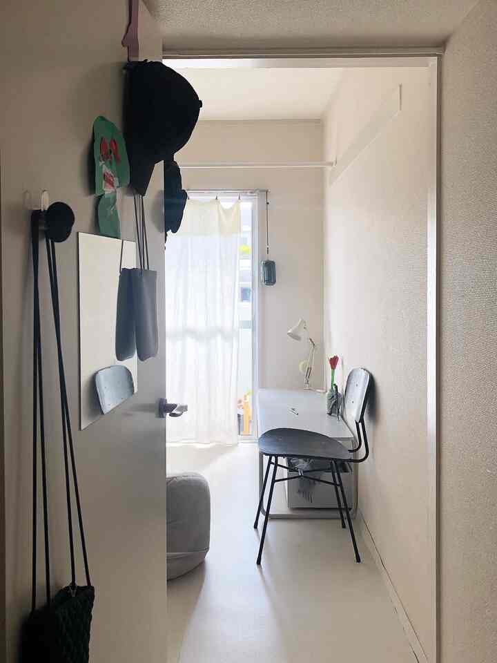 白とベージュのトーンの狭い部屋、シンプルなデスクと椅子、明るいカーテンが配置されたミニマルな空間